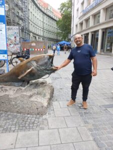 Fotografie de referință a unui bărbat cu ochelari și tricou albastru, lângă o sculptură de pește.